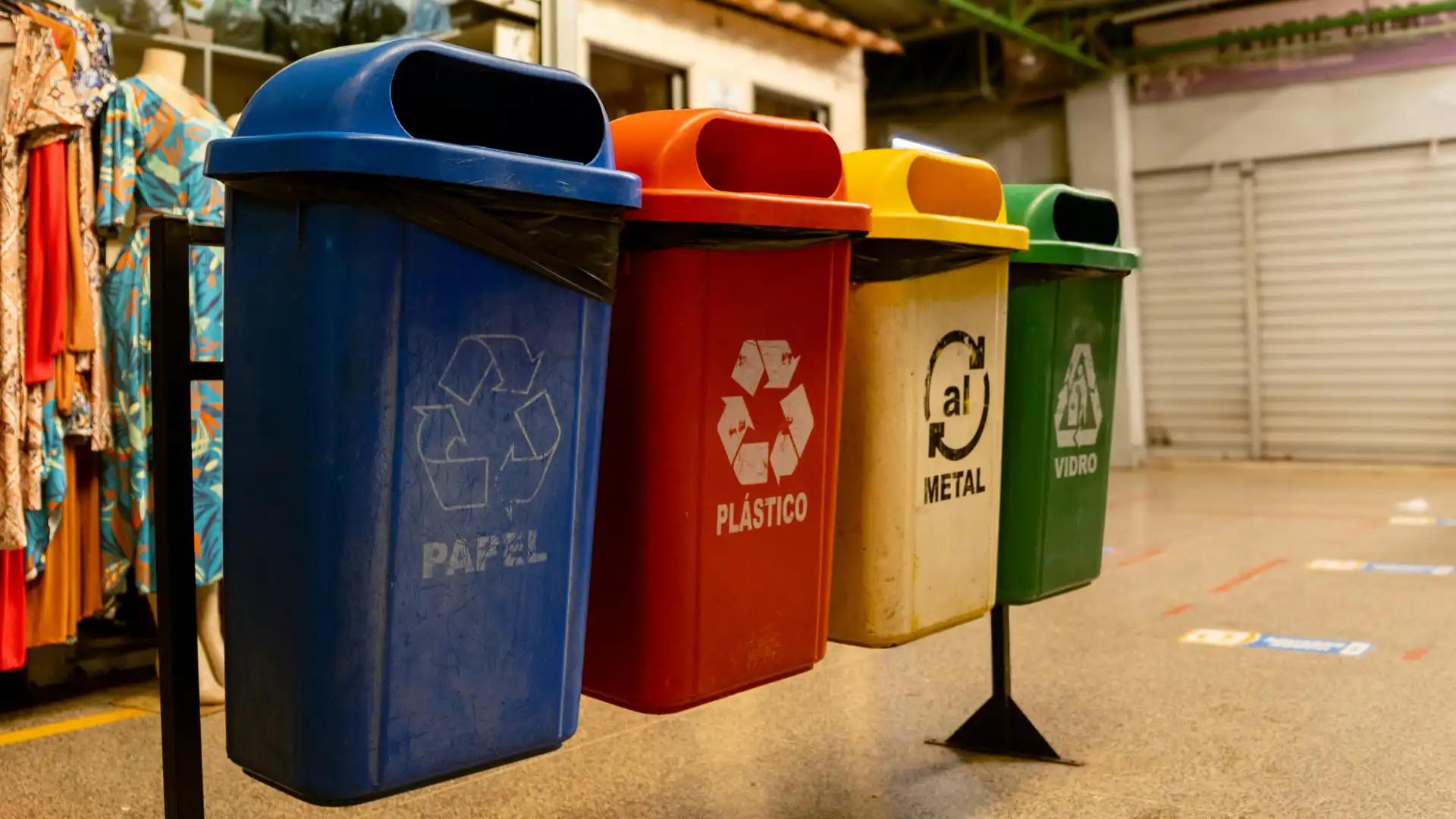 Imagem de cestas de lixo coloridas em uma rua, incluindo recipientes para papel, plástico, metal e vidro, utilizadas para separação de resíduos e reciclagem.