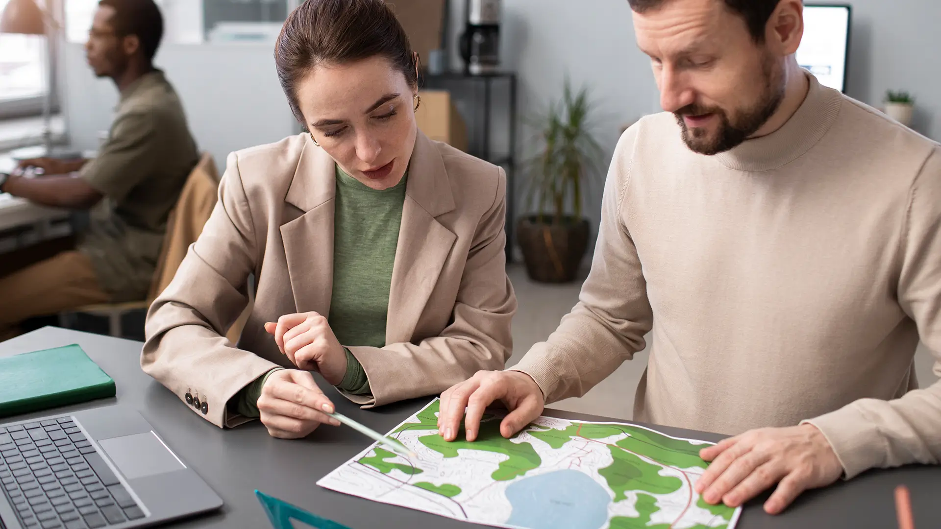 Engenheiros analisando projeto de planejamento de área verde, com mapa e laptop na mesa, em escritório moderno e bem iluminado.