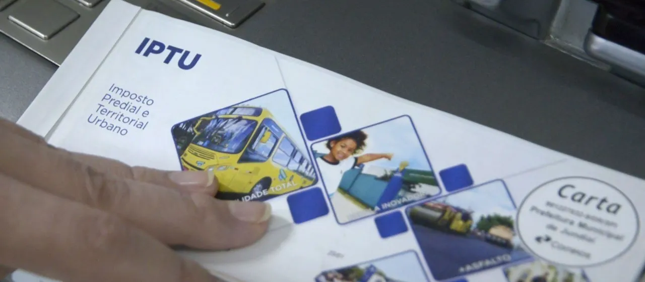 Folha de papel com informações do IPTU, incluindo detalhes sobre o Imposto Predial e Territorial Urbano, com uma pessoa segurando o documento, visualizado em um ambiente de escritório.